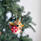 A handmade felt hedgehog ornament is nestled among the vibrant green branches of a Christmas tree. This charming wool felt hedgehog features rainbow-colored quills in blue, yellow, orange, and green, and holds a red-and-white spotted mushroom. A natural jute string loop is attached for easy hanging, adding a cozy touch.