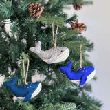 A felt humpback whale ornament hangs on a Christmas tree adorned with pine cones, set against a soft, neutral background. This unique wool felt humpback whale ornament features a light gray body and a white underbelly, with tiny white embroidered details that highlight its needle-felted, hand-stitched texture. It has a small tail and flippers, with a jute string attached for hanging. The handcrafted ornament adds a cozy, festive charm.