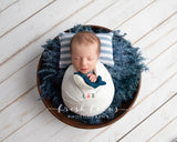 A handmade felt humpback whale Christmas ornament in dark blue and white rests in the arms of a swaddled newborn. This handcrafted needle-felted wool ornament features a blue humpback whale design with subtle white details, meticulously hand-stitched.