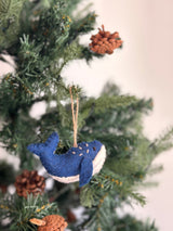 A felt humpback whale ornament is displayed on a Christmas tree, nestled among evergreen branches and pine cones. The gray whale features a white underbelly and textured detailing on its back. Crafted from organic wool with needle-felted and hand-stitched features, a jute string loop at the top allows for easy hanging as a delightful Christmas tree ornament.
