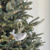 A needle felted gray humpback whale felt Christmas ornament hangs from a jute string on a brightly lit Christmas tree. The handcrafted whale features a lighter gray lower jaw and a dark eye, nestled among green needles and warm white lights, evoking a cozy and festive mood.