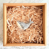 A wool felt humpback whale ornament rests in a brown cardboard box, nestled among light brown paper shred. This gray and white felted ornament features a rounded body, a distinct tail, and a jute string attached for hanging.
