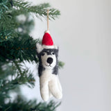 A charming felt husky ornament with a red Christmas hat hangs gracefully on a vibrant green Christmas tree, set against a soft neutral wall. This unique Christmas ornament is crafted from white and gray felt, featuring a round black nose, a sweet smile, and a white pompom. A jute string secures the handcrafted felt ornament.