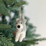 A handcrafted husky felt ornament hangs gracefully from a lush green Christmas tree branch. This unique felt ornament features a blend of gray and white felt, accurately capturing the husky's coat, complete with pointed ears, a black nose, and a bushy tail. It is suspended by a jute string, adding a touch of handmade elegance to the festive decor.
