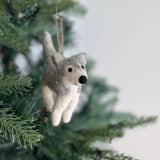 A handcrafted felt husky ornament hangs from a Christmas tree branch, creating a festive and joyful atmosphere. This unique Christmas ornament showcases a blend of gray and white felt, capturing the husky's distinctive markings with small black eyes and nose. Its cozy felt texture contributes to a handcrafted feel, suspended by a jute string.