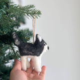 A handmade felt husky ornament is held by a female hand, suspended from a Christmas tree branch. This charming felt husky ornament features a distinctive black and white pattern, with a black body, bushy tail, and white accents on its face, ears, and legs. Its textured surface provides a soft, plush appearance, perfect for adding a whimsical touch to holiday decor.