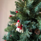 A charming wool felt Jack Russell dog Christmas ornament, adorned with a red hat and scarf, hangs on a festive green Christmas tree. This handmade felt ornament, crafted from white and brown organic wool, features a black nose, a cheerful smile, and floppy brown ears. The vibrant tree is decorated with red berries and brown pine cones.
