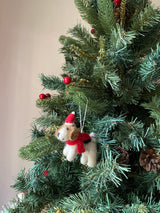 A cheerful wool felt Jack Russell dog ornament with a Christmas hat hangs prominently on a green Christmas tree. The adorable 3D-designed ornament features a red scarf and a festive red and white hat, with a white string for hanging. Red berries and brown pine cones decorate the tree, adding to the festive mood.