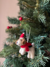 A wool felt Jack Russell dog Christmas ornament hangs from a festive Christmas tree branch adorned with green needles and red berries. The cream-colored dog features brown markings on its head, small black eyes, and wears a red Christmas hat with a white pompom and a matching scarf. A thin white string for hanging is attached to the hat.