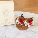 Two wool felt Jack Russell dog ornaments are displayed on a woven mat set on a white marble table, with a book featuring a botanical rose illustration in the background. These handmade ornaments have brown and white bodies with floppy brown ears. Each features a festive red cotton string around its neck and a metal ring on its back, highlighting their artisanal charm.