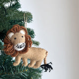 A handcrafted felt lion ornament hangs from a green Christmas tree branch against a soft gray background. The felt lion features a tan body with a fluffy brown mane, white inner ears, and a black yarn tail. Its face is detailed with black eyes, a stitched nose, and whiskers, and it has a jute string for hanging.