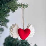 A handcrafted, needle-felted Love with Wing felt ornament hangs from a Christmas tree branch against a soft backdrop. This 3D Christmas tree ornament features a white wing base with a striking red 'love' motif beautifully needle-felted onto it. The wing has layered, scalloped tiers creating a feathered effect and is suspended by a delicate jute string.