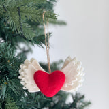 A needle felted Love with Wing ornament hangs on a Christmas tree, creating a festive and heartwarming atmosphere. This 3D ornament features a striking red 'love' motif beautifully needle-felted onto a soft white wing with intricately textured surfaces. It is suspended by a jute string, adding a rustic touch to the visually striking piece.