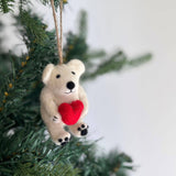 A handcrafted Mini Polar Bear Holding Heart felt ornament hangs gracefully on a Christmas tree. This charming white wool felt polar bear features a black nose, tiny black eyes, and feet, holding a bright red heart. A jute string allows this unique Christmas ornament to add a joyful, festive touch to any tree.