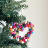 A wool felt Mini Pompom Heart Wreath ornament hangs from a Christmas tree branch against a soft white background. This petite, heart-shaped felt ornament is adorned with vibrant pink, red, white, blue, and orange mini pompoms, with a jute string loop at the top for hanging.