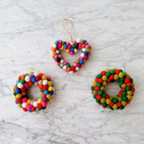 A wool felt mini pompom heart wreath ornament hangs suspended by a jute string against a light gray marble backdrop. This vibrant, handcrafted felt ornament is densely packed with mini pompoms in shades of red, green, blue, yellow, white, and pink, creating a charming textured surface.