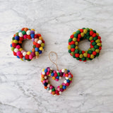 A vibrant, multicolored wool felt mini pompom heart wreath ornament rests brightly on a white marble surface. This handcrafted felt ornament features a heart shape adorned with numerous red, pink, green, yellow, blue, and white mini pompoms. A jute string is attached for easy hanging, highlighting its joyful and playful nature.