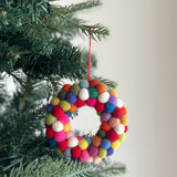 A colorful mini pompom heart-shaped wreath felt ornament hangs from a pine branch on a Christmas tree, secured by a red cotton string. This wool felt ornament features an array of mini pompoms in red, white, orange, blue, green, yellow, and pink, forming a textured, handcrafted heart.