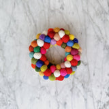 A handcrafted wool felt mini pompom wreath ornament rests on a light gray marble background. This petite heart-shaped Christmas ornament features a dense arrangement of colorful felt pompoms in shades of red, white, green, yellow, blue, orange, and purple, offering a soft and tactile, joyful aesthetic.