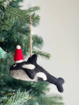 A playful needle felted orca ornament with a Christmas hat hangs on a vibrant green Christmas tree. This unique Christmas ornament is crafted from soft wool felt in a harmonious blend of gray, black, and white, featuring distinct white markings. The charming orca is adorned with a red and white Santa hat and includes a jute string for easy hanging.
