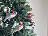 Several handcrafted felt orca ornaments hang festively on a Christmas tree, nestled among green needles and natural pine cones. These unique felt orca ornaments, in varied shades of grey, brown, and black, are suspended by a jute string from the tree, each adorning a small red and white Santa hat, creating a warm and joyful scene.
