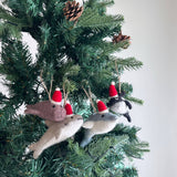 Felt orca ornaments, each wearing a red Christmas hat, hang joyfully on a green Christmas tree adorned with pine cones. These playful handmade felt ornaments feature a variety of colors, including white, gray, light blue, dark gray, and purple. Each detailed orca ornament has a tail, small side fins, and a distinct white belly, completing the festive look against a plain white wall.