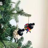 Two charming Pompom Ball Mini Sheep felt ornaments hang on a Christmas tree branch, adding a joyful festive touch. One is a white sheep with a black face and a golden bell. The other is a colorful sheep with a black face and a body of red, blue, yellow, and green felt pompom balls, also with a golden bell.