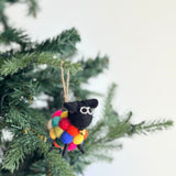 A festive felt mini sheep ornament, with a body made of colorful pompom balls, hangs joyfully on a Christmas tree. This unique Christmas tree ornament features a black head with white eyes and a golden bell attached around its neck, ready for hanging by a jute string.