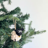 A Felt Pompom Ball Mini Sheep Christmas ornament hangs on a Christmas tree branch. This charming wool felt sheep ornament features a cream-colored, pompom-textured body with a black head and ears. Its round eyes are white with black pupils. The handcrafted ornament is suspended by a jute string.