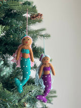 Two vibrant handcrafted felt Pride Mermaid ornaments hang gracefully on a Christmas tree with vivid green needles. One mermaid features light brown skin, pink hair, a turquoise top, and a matching tail adorned with white dots, suspended by a pink string. The second mermaid has light brown skin, light blue hair, a purple top, and a coordinating tail with white dots, hanging from a light blue string. These unique Christmas ornaments embody self-expression and radiant joy.