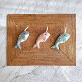 A set of three handmade felt narwhal ornaments rests on a rectangular woven Bali rattan placemat, displayed against a light marble surface. Each cheerful narwhal ornament features a unique color: pink, baby blue, or ocean blue, with white spots on its back and a white horn. The rustic mat enhances their handcrafted charm.