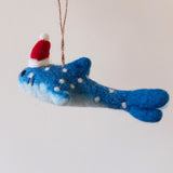 A smiley whale shark felt ornament is suspended by a brown jute string against a plain background. This unique Christmas felt ornament features a light to dark blue gradient body. It is adorned with evenly spaced white dots and black stitched eyes, creating a cute, happy expression.