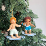 Two cheerful felt surfer boy ornaments hang against a green Christmas tree with pinecones, bringing a summery vibe. One tan-skinned boy with orange curly hair and mint green shorts smiles while riding a light blue surfboard. The other, a brown-skinned boy with brown curly hair, blue shorts, and a blue bracelet, also smiles while on a matching surfboard. Both handmade felt ornaments have red stitched mouths and white trim on their surfboards.