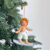 A handcrafted felt surfer boy ornament hangs on a Christmas tree, adding a joyful, festive touch. The cheerful surfer boy features orange curly hair, tan skin, and light green shorts with white trim. He rides a light blue surfboard with white detailing, ready to hang from an orange string.