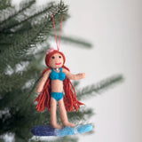 A playful handcrafted felt surfer girl ornament hangs from a green Christmas tree branch. This unique felt ornament depicts a girl with long red hair, a flower on her head, a red smile, and a pearl choker necklace. She wears a blue bikini while riding a blue and light blue surfboard, with a red cotton string attached for hanging.