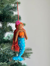 A whimsical felt tropical mermaid ornament dangles gracefully from a green Christmas tree branch. The handmade felt mermaid features rich brown skin, flowing sunset-toned yarn hair, an orange top, and a bright blue tail adorned with white pearly details.