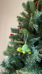 A handmade organic wool felt turtle ornament hangs on a festive Christmas tree, bringing nature-inspired charm to the setting. The 3D felt turtle features a light green body and a darker green shell, with a slightly raised head and small round eyes. A jute string is attached for hanging this charming, hand-stitched ornament, visible amidst red berries and artificial pine needles.