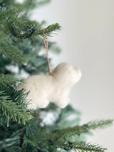 A white Maltese dog felt ornament hangs on a festive Christmas tree. This handcrafted felt ornament has a fluffy white texture, mimicking the soft fur of a Maltese dog. It hangs from a simple jute string, looped over a green pine needle branch, amidst the tree's warm glowing lights.
