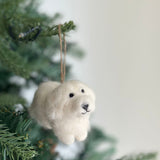 A handcrafted felt Maltese dog ornament hangs from a lush green Christmas tree branch, evoking cozy holiday warmth. This white felt Maltese dog, suspended by a jute string, features small black eyes and a stitched black nose and mouth, adding a personal and festive touch to holiday decor.