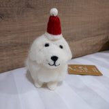A handcrafted white Maltese dog felt ornament stands on a subtly striped white surface, against a soft-focus wood-paneled wall. A small tag rests beside this charming felt ornament, which is shaped like a white Maltese dog with dark eyes and nose, and a sweet expression. It wears a jaunty red felt Christmas hat topped with a white pompom, giving it a soft texture and whimsical appeal as a Christmas decoration.