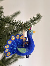 A deep blue wool felt peacock ornament hangs from a jute thread against festive evergreen branches and a white wall. This handmade felt peacock ornament features a textured body, a small yellow beak, and colorful plumage in red, orange, and green. Its tail is adorned with teardrop-shaped patterns of gold, white, and blue, perfect for hanging on a Christmas tree.