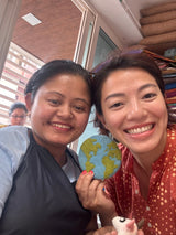 A round felt Planet Earth coaster is held by two smiling women in a bright, cheerful setting. The organic lamb wool felt coaster features green continents against a blue ocean backdrop, with visible stitching highlighting its ethically handcrafted quality. These unique, eco-friendly coasters evoke global awareness.