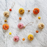 A felt pumpkin garland is displayed on a marbled white surface, presenting an autumnal mood. The garland features eight autumn-colored, felted wool pumpkins, strung together with a thin jute thread visible between each one.