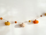A vibrant felt pumpkin garland hangs gracefully against a clean white wall. The garland features multiple wool felt pumpkins in warm shades of orange, yellow, and beige. Each pumpkin has a textured, ridged surface, strung together with a natural jute thread, creating a rustic and inviting display.