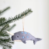 A gray felt narwhal ornament hangs from a lush green pine branch against a soft gray backdrop. This handmade felt narwhal features a white horn and belly, light blue felt circles, and a small black eye. Its textured material gives it a cozy feel, suspended by a jute string, adding a unique, handcrafted touch to holiday decor.
