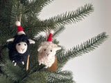 Two cozy wool felt sheep ornaments with Christmas hats, one black and one white, are nestled among vibrant green pine needles. Each ornament features fuzzy wool fur, a cream-colored face with a stitched 'Y' mouth, and a small red and white Santa hat. The white sheep also wears a colorful scarf, both ready to hang with a hemp string loop, evoking a festive mood.