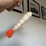 A felt shrimp ornament, suspended by a jute string, is gently held by a female hand against a bright backdrop with framed pictures. The handcrafted felt shrimp ornament is predominantly white with light pink stripes around its body and features a vibrant orange tail, offering a unique, sea-themed touch.