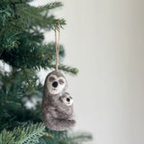 A wool felt sloth ornament, depicting a gray mother sloth with a white face, black nose, and closed eyes tenderly embracing her smaller baby, hangs from a green Christmas tree branch. This handcrafted felt Christmas tree ornament, made from textured gray wool felt, features a jute string for hanging and evokes a sense of warmth and familial love.