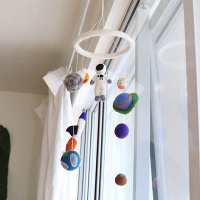 A felt spaceman baby mobile hangs in front of a bright window with sheer white curtains, creating a whimsical, dreamy scene. The mobile features a top ring suspending needle-felted 3D figures by thin white strings. These include a white spaceman, a blue and white rocket with orange flames, a grey asteroid, and colorful planets in shades of blue, purple, orange, and brown.