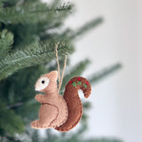 A light brown wool felt squirrel ornament hangs from a green Christmas tree branch. Its darker bushy tail is adorned with delicate hand embroidery featuring red berries and green sprigs. This handcrafted felt ornament adds a cozy, rustic woodland touch to holiday decor.